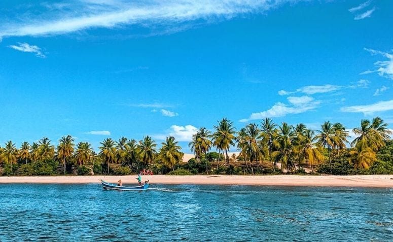 Vista panorâmica de Mangue Seco com praia de areia clara, coqueirais, barco de pescadores e águas calmas do Rio Real, na divisa entre Sergipe e Bahia.