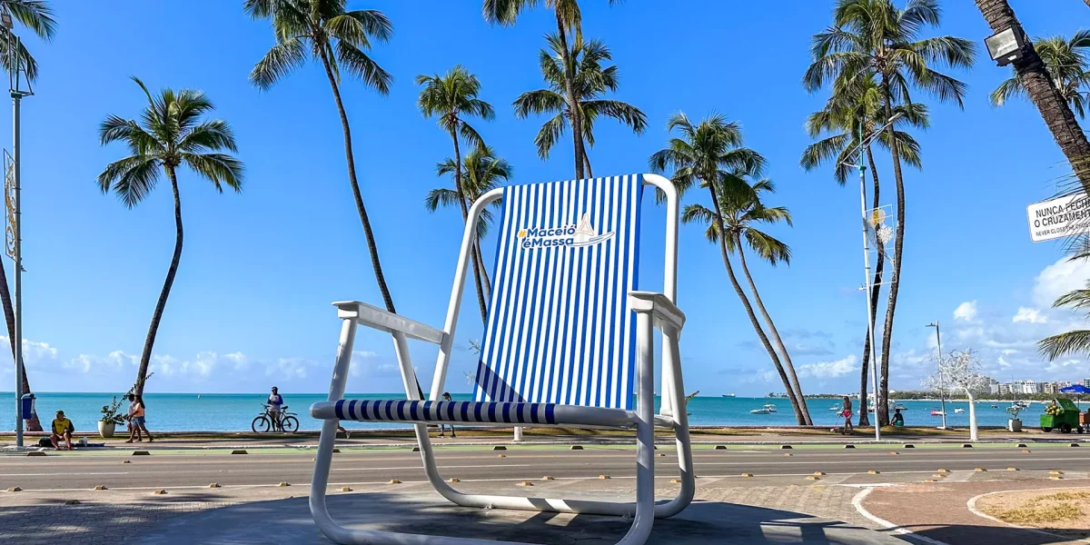 Cadeira gigante Maceió é Massa na orla de Maceió, Alagoas, com coqueiros, mar azul, céu ensolarado e paisagem turística do litoral nordestino.