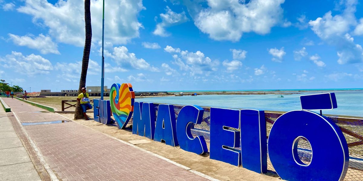 Letreiro Eu amo Maceió na orla de Maceió, Alagoas, com mar azul, céu ensolarado e paisagem turística do litoral nordestino.