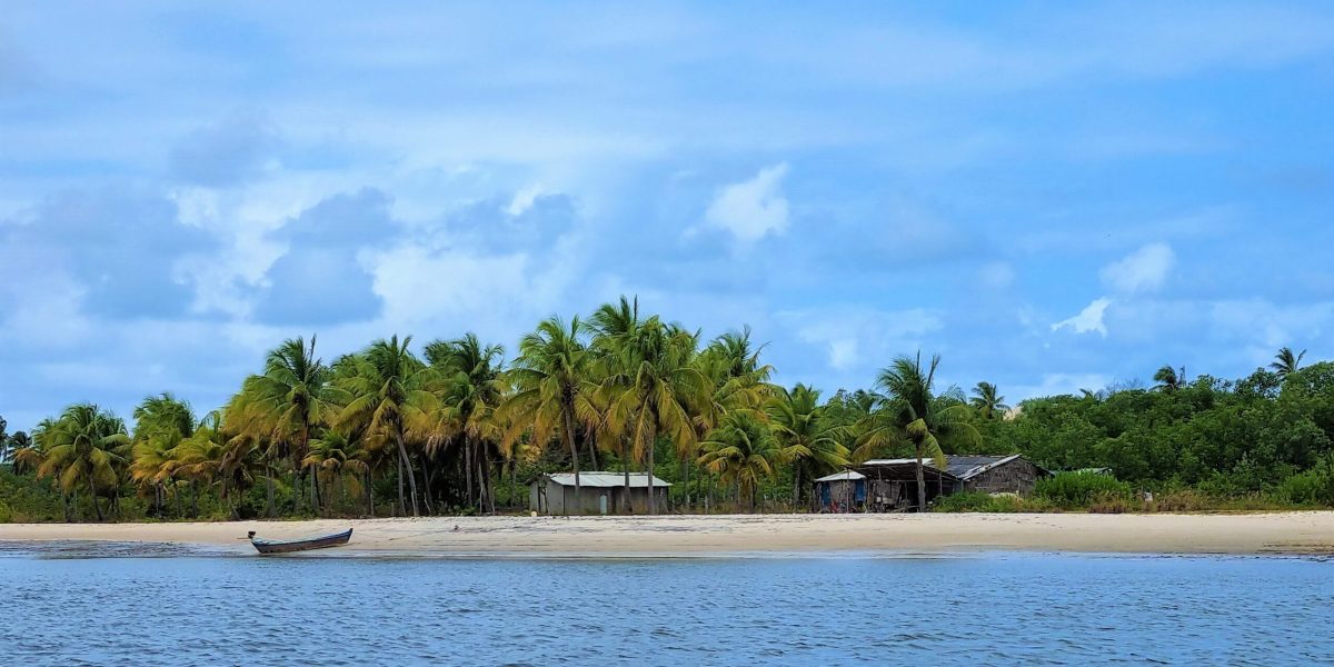 Paisagem de Mangue Seco na Bahia com coqueiros, praia de areia clara, barco à beira da água e cenário natural do litoral nordestino.