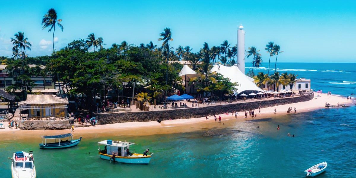 Vista aérea da Praia do Forte na Bahia com farol, barcos coloridos, mar cristalino, coqueiros e faixa de areia no litoral norte baiano.